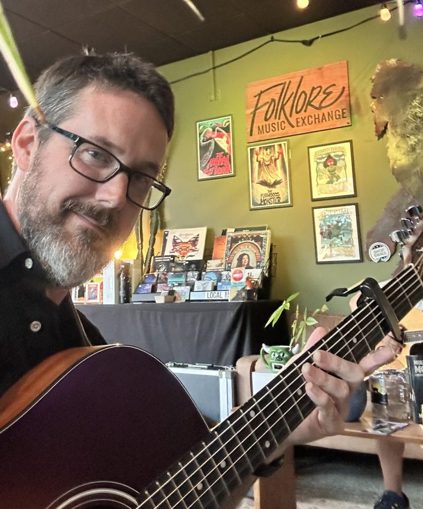 A man with glasses and a beard is holding a purple guitar in a cozy music shop adorned with posters and a sign that reads 'Folklore Music Exchange.'